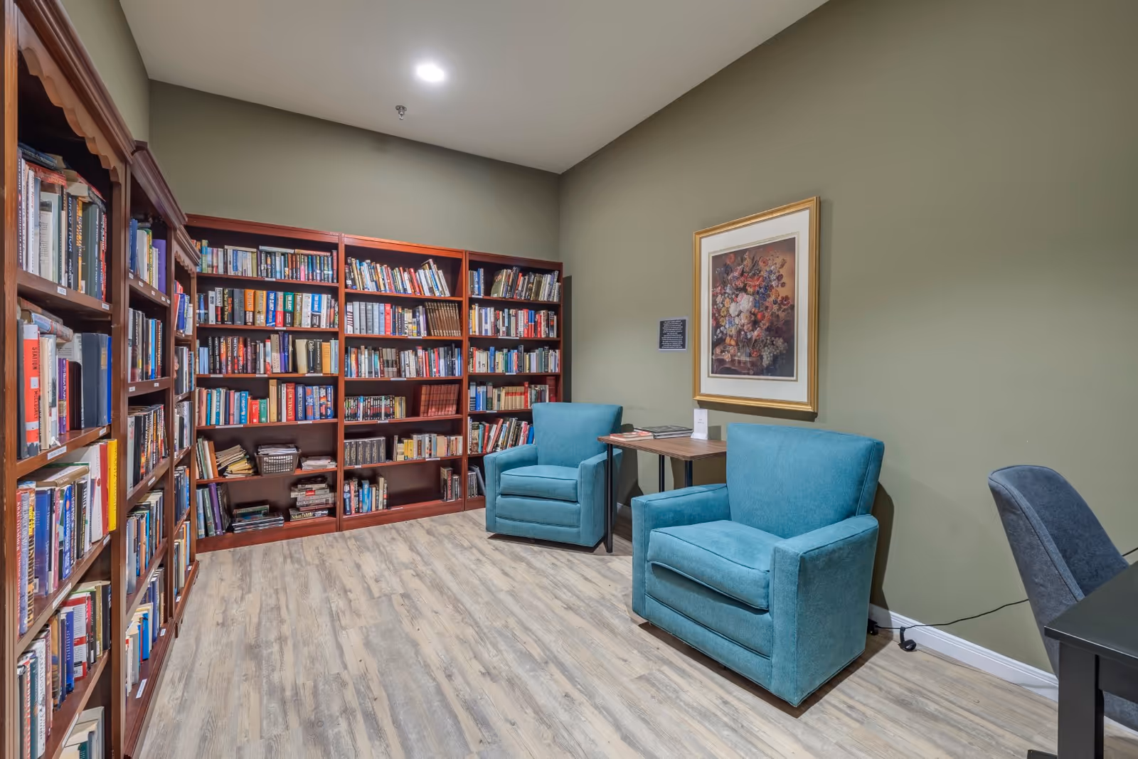Cozy library corner with tall bookshelves, teal armchairs, a table, and framed art.