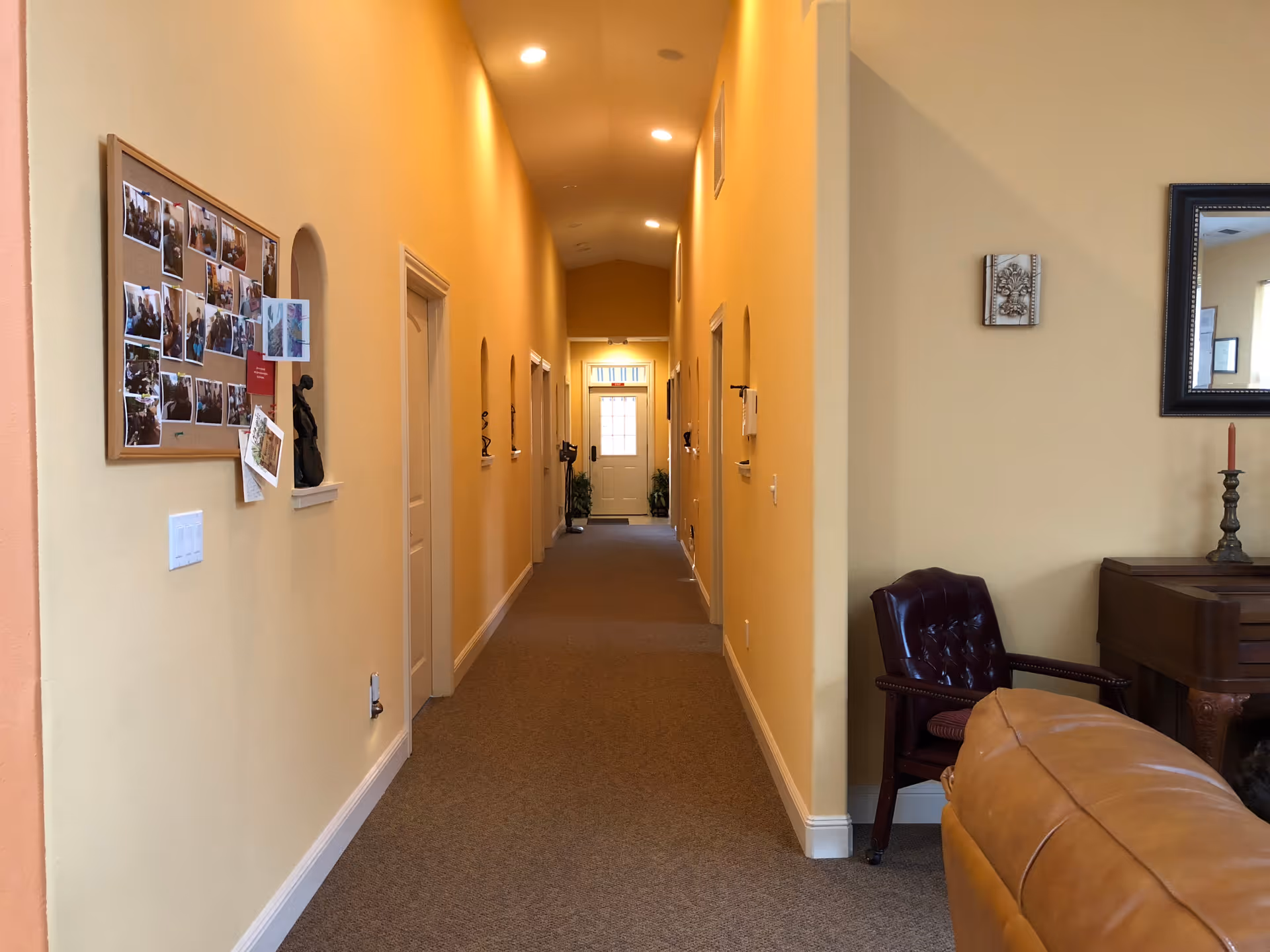 A long, well-lit hallway with beige walls and carpeted floor in a senior care facility. On the left wall, there is a bulletin board with various photos and papers pinned to it. The hallway has several doorways and small arched openings along the walls. At the far end, a door with a window is visible. On the right side, part of a living area is visible with a brown leather chair, a wooden piano, a framed mirror, and a candle holder.