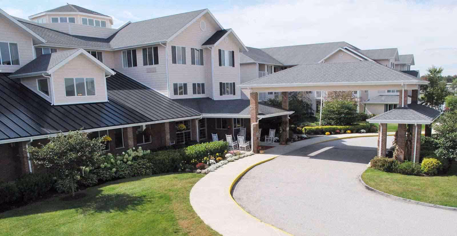 Exterior view of a senior living facility with a covered entrance driveway, multiple windows, and a landscaped garden with green grass and shrubs.