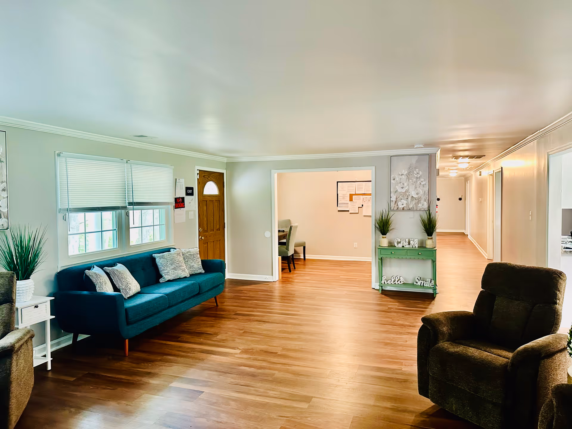 A bright and spacious living room area in Ascension Assisted Living Facility featuring a teal sofa with decorative pillows, two brown armchairs, a small white side table with a plant, and a green console table with plants and decorative items. The room has wooden flooring, light-colored walls, and a wooden door with a window. There is an open doorway leading to another room with chairs and a bulletin board.