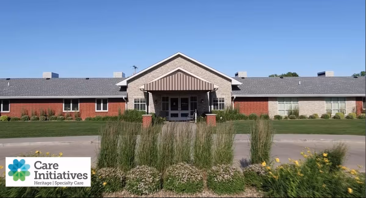 Single-story brick senior care building with a central covered entrance and landscaped lawn under a clear sky.
