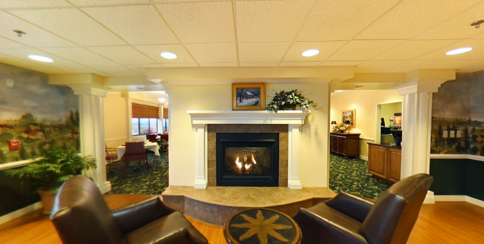 A cozy sitting area with two brown leather chairs facing a lit fireplace with a white mantel. Above the mantel is a framed painting and a plant. The room has a patterned carpet and walls with murals, and there are dining tables visible through the archways on either side of the fireplace.