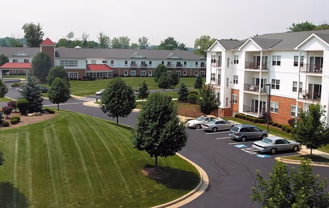 Aerial view of a senior living campus with multi-story residential buildings, parked cars, and landscaped lawns and trees.