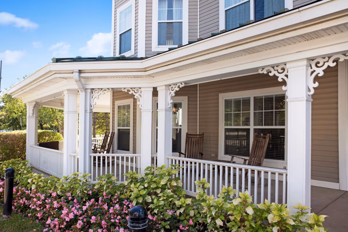 Front porch of a residential building with white columns, rocking chairs, and flower beds.