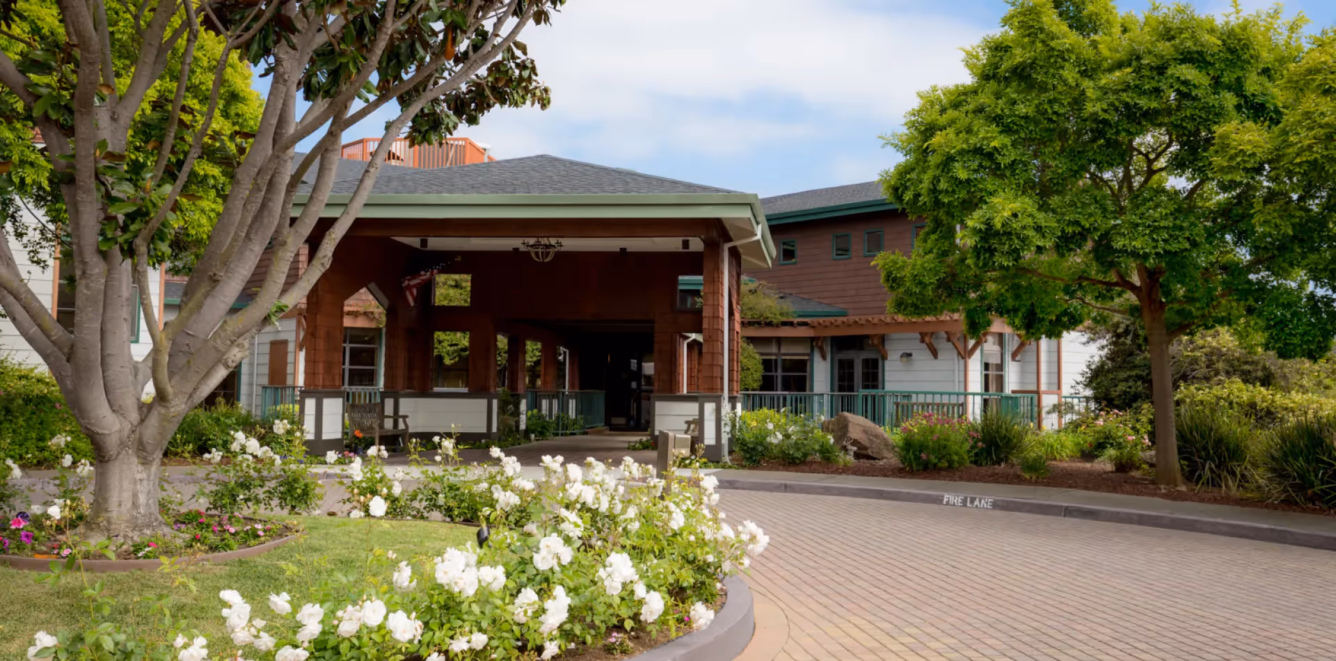Front entrance of a senior living building with a covered porte-cochere, landscaped flowerbeds and trees.