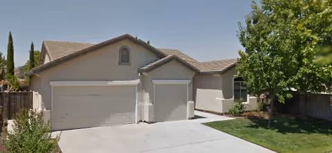 Single-story beige senior living facility with two garage doors, driveway, and a tree on the right.