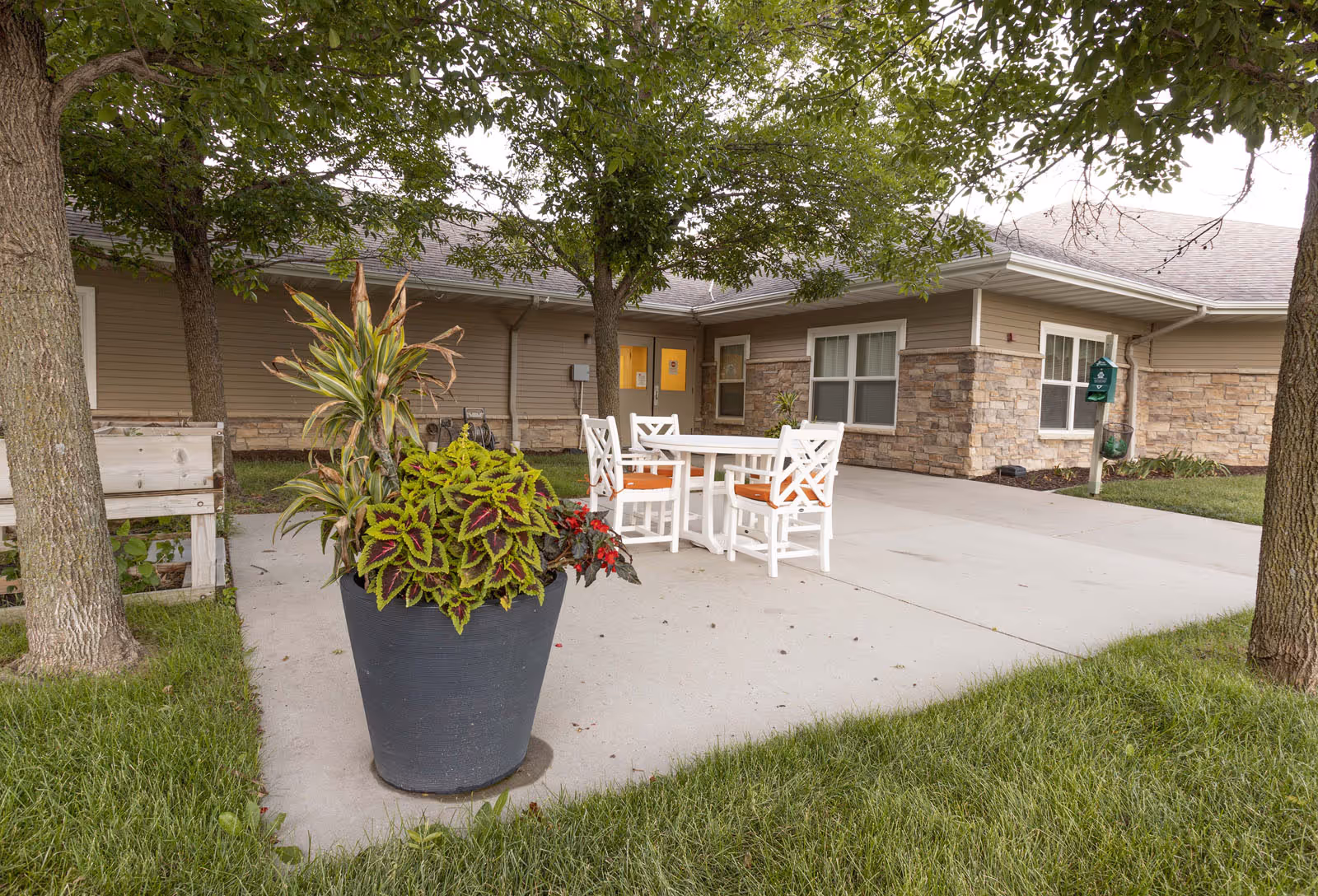 Outdoor patio area at Independence Village Of Waukee featuring a large planter with various green and red plants, white table with four white chairs with orange cushions, surrounded by trees and grass, adjacent to a building with stone and beige siding.