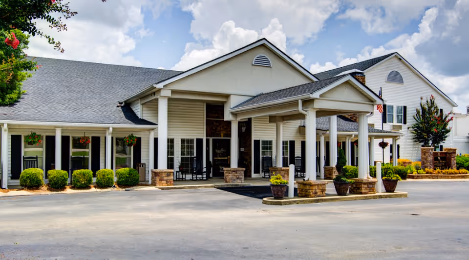 Front entrance of a white senior living facility with a covered porte-cochere, columns, rocking chairs, and landscaped shrubs.