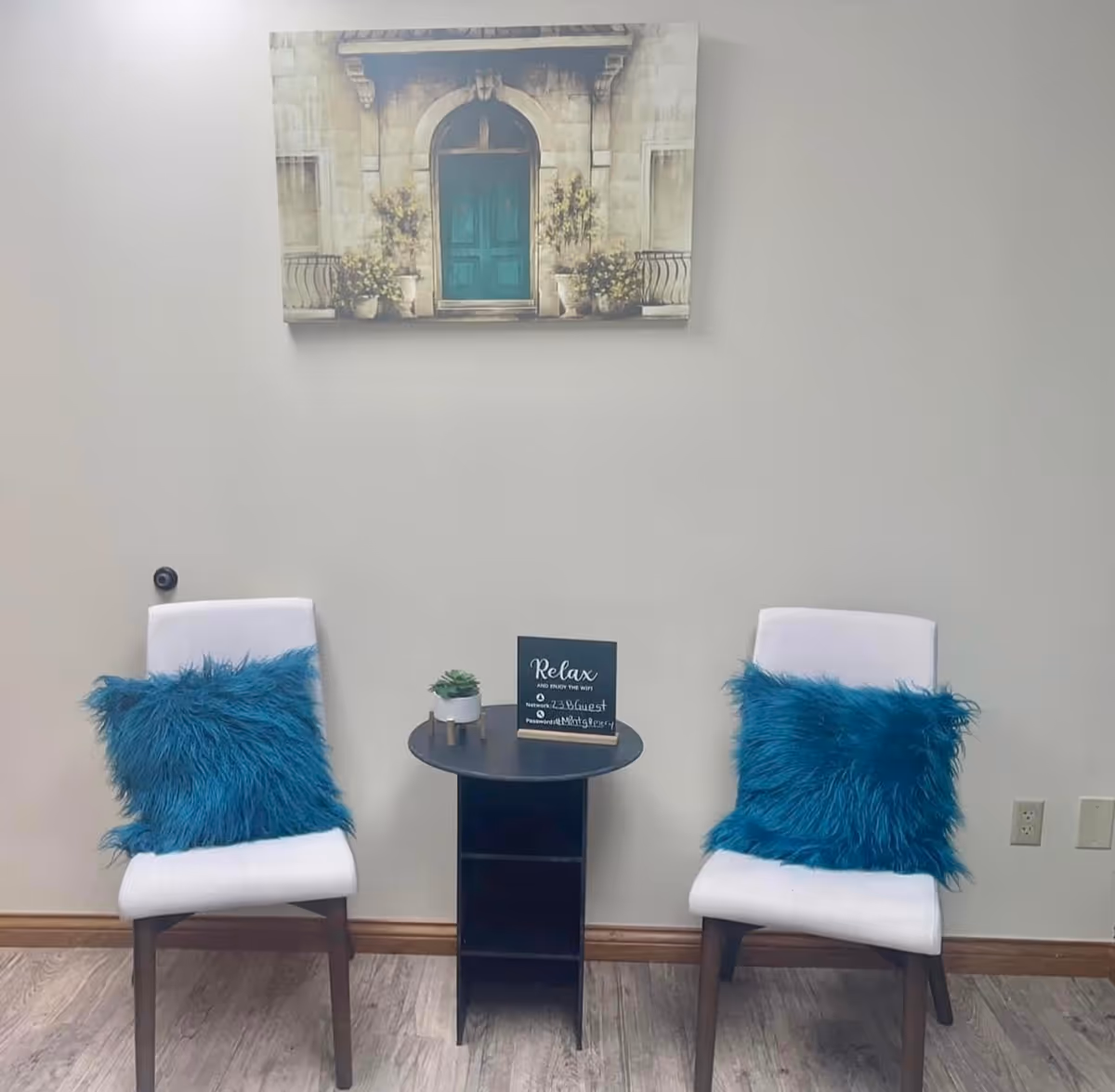 Two white chairs with blue cushions flank a small black table beneath wall art.