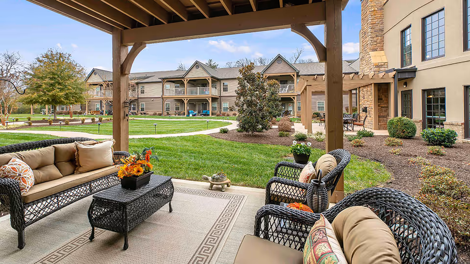 Outdoor covered patio area with wicker seating including a sofa and chairs with cushions and decorative pillows, a coffee table with a flower arrangement, and a small turtle statue on the ground. The patio overlooks a well-maintained grassy courtyard with shrubs, trees, and a two-story building with balconies in the background under a partly cloudy sky.