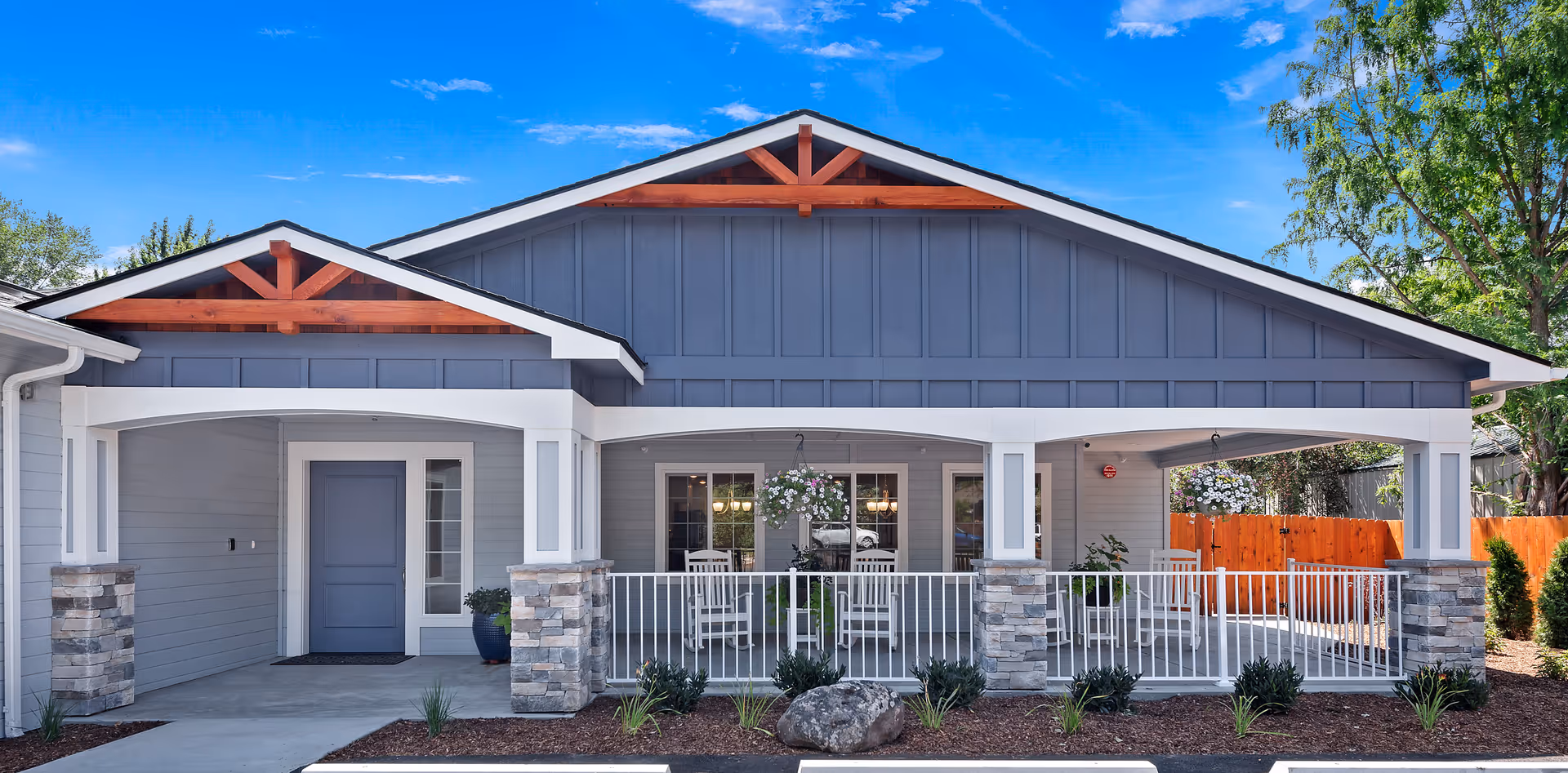 Front exterior view of a single-story building with a covered porch supported by white columns with stone bases. The building has gray siding with blue accents and wooden decorative elements under the roof peaks. There are hanging flower baskets on the porch, white rocking chairs, and a wooden fence in the background under a bright blue sky.