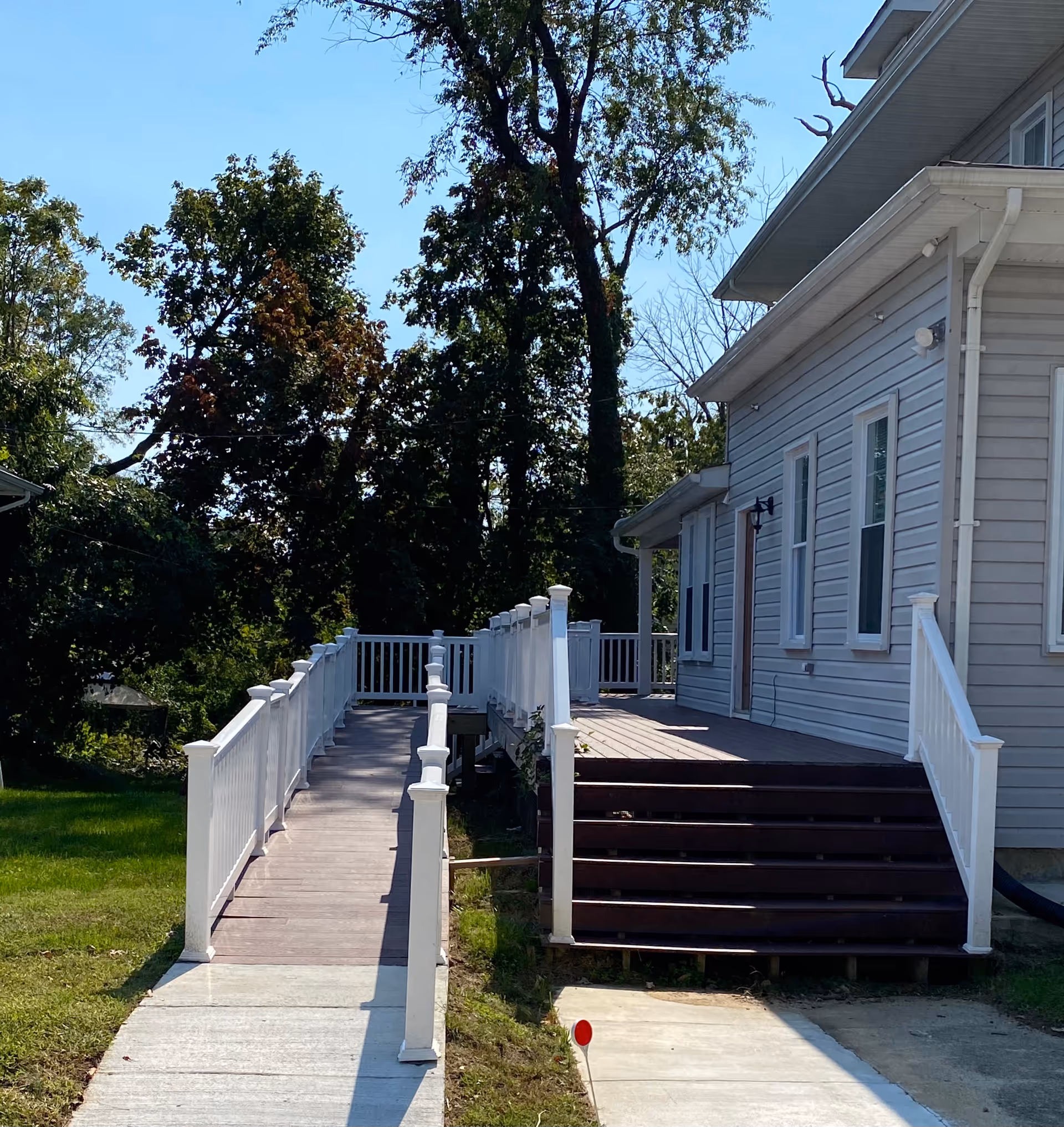 Exterior view of a light-gray siding building with a wooden ramp and steps leading to a raised porch, with trees in the background.