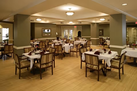 A spacious senior living dining room with many tables set with white tablecloths.