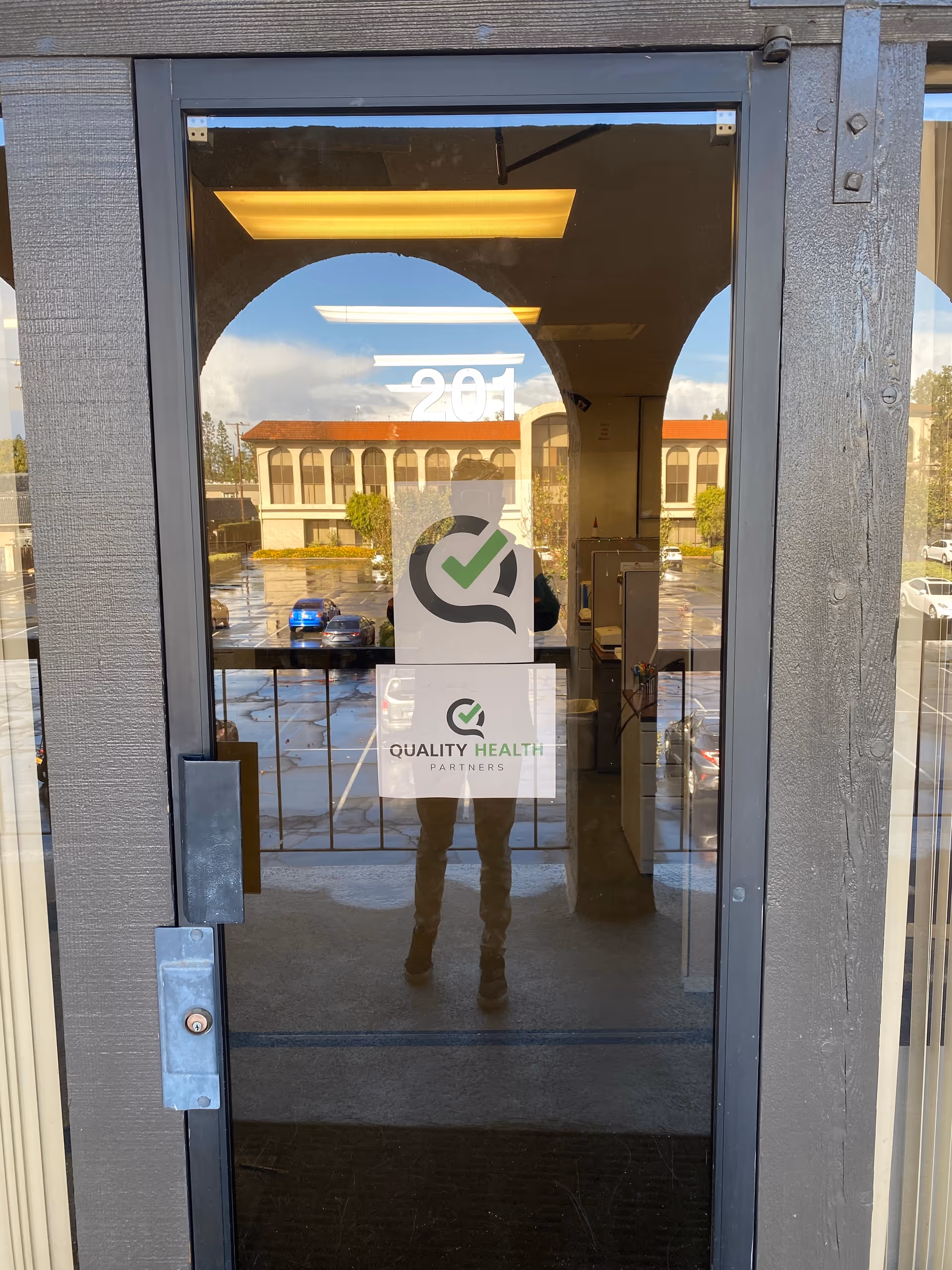 Glass entrance door shows a Quality Health Partners sign and reflected parking lot.