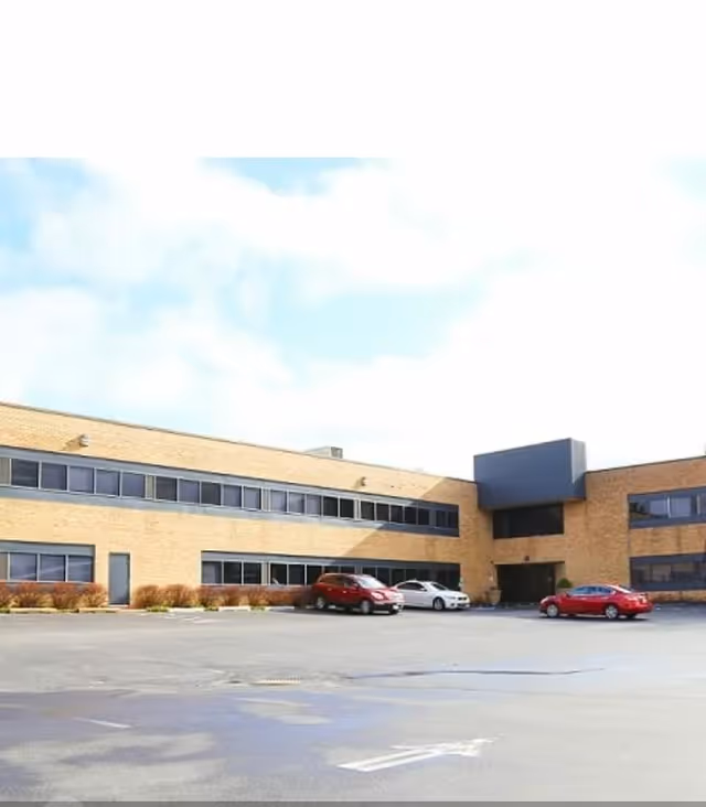 Two-story brick facility with a blue entrance, a large parking lot, and several cars.