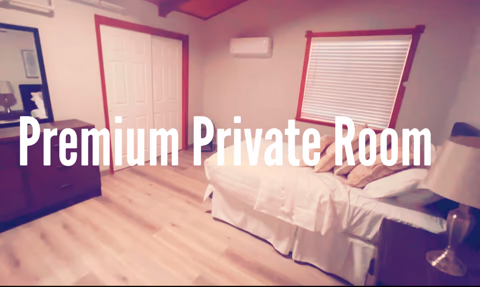 Private bedroom in a care facility featuring a bed, dresser, window blinds, and wall AC.