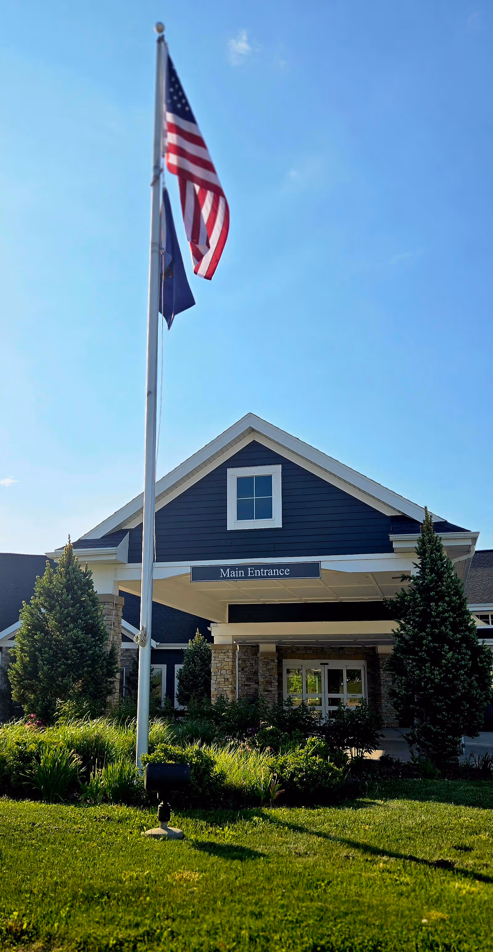 Front entrance of a blue building with a peaked roof, flagpole, and landscaping.