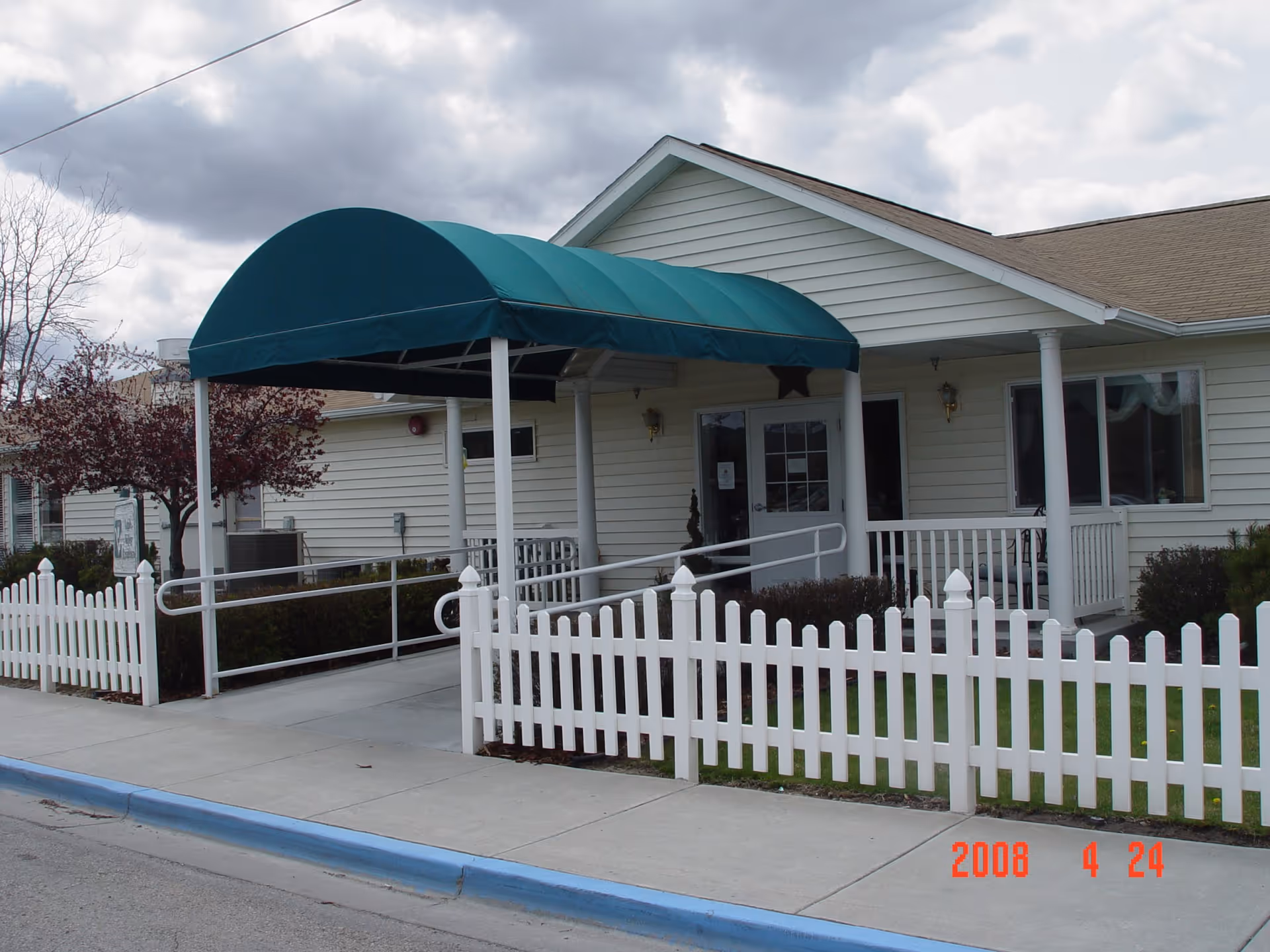 Exterior view of a single-story building with white siding and a green awning over the entrance. There is a white picket fence surrounding a small grassy area and a concrete sidewalk leading to the entrance with a wheelchair ramp. The sky is cloudy.