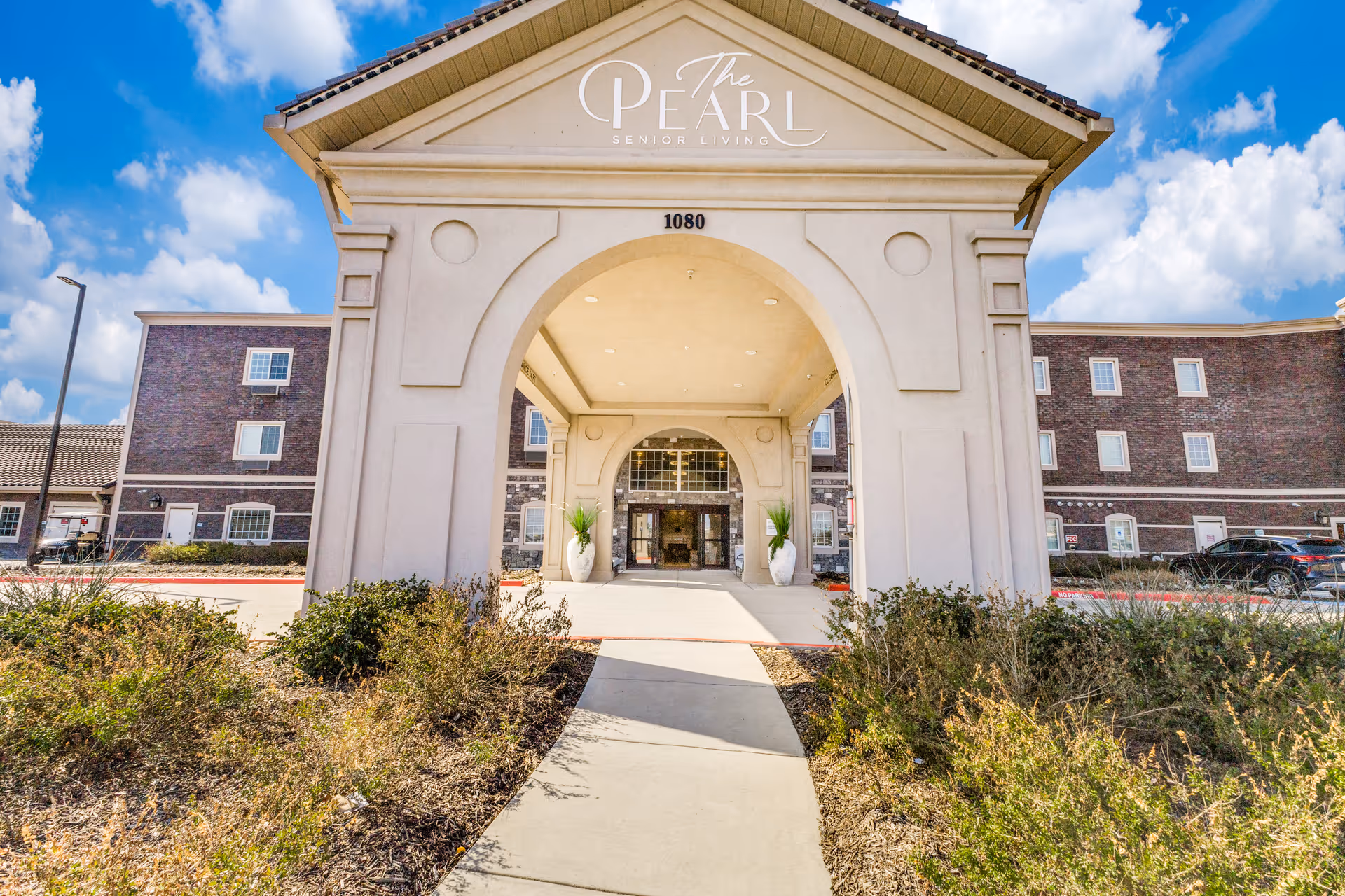 Front entrance of The Pearl Senior Living building with a large arched portico and walkway leading to the doors.