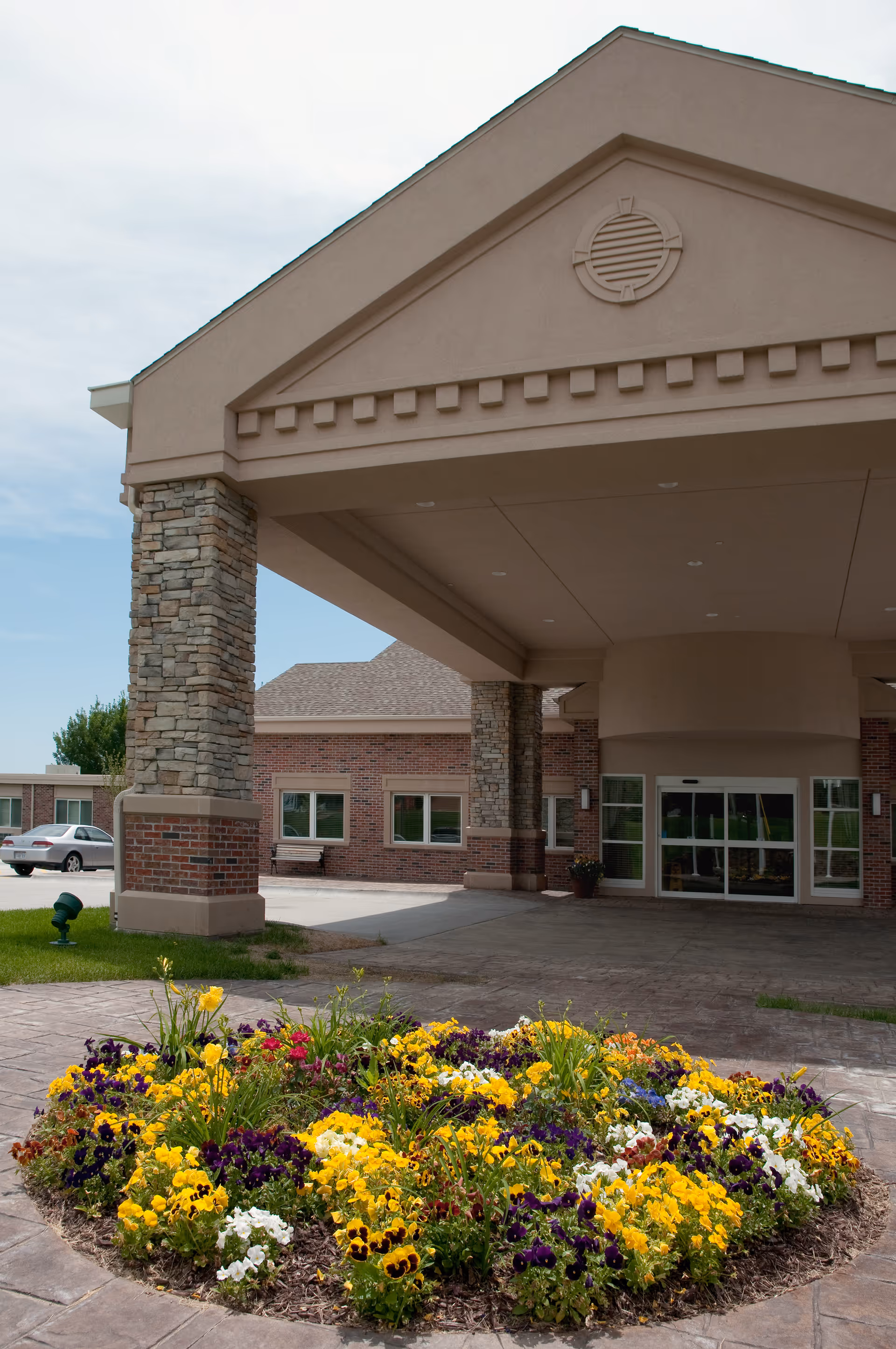 Front entrance of a brick senior living facility with a covered porte-cochère supported by stone columns and a circular flower bed in the foreground.
