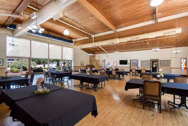 Spacious dining room with wooden floors and ceiling, several tables covered with black tablecloths, each adorned with small floral centerpieces. Large windows on one side let in natural light and offer a view of greenery outside. The room has multiple chairs around the tables, ceiling lights, and a stone fireplace in the back corner.