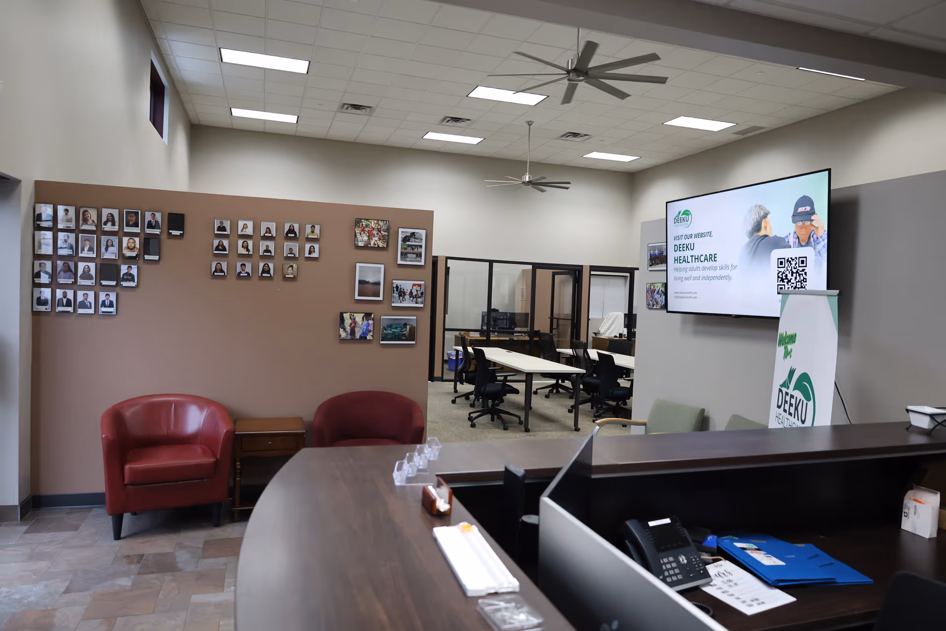 Lobby area of Deeku Healthcare with curved reception desk, two red chairs, wall photos, and screen.