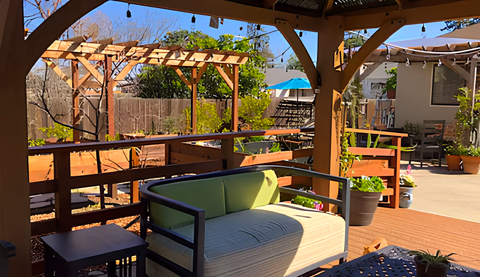Outdoor covered patio area with a cushioned bench and small tables, surrounded by wooden railings and planters with greenery. In the background, there are wooden pergolas, potted plants, and a blue umbrella providing shade over a seating area.