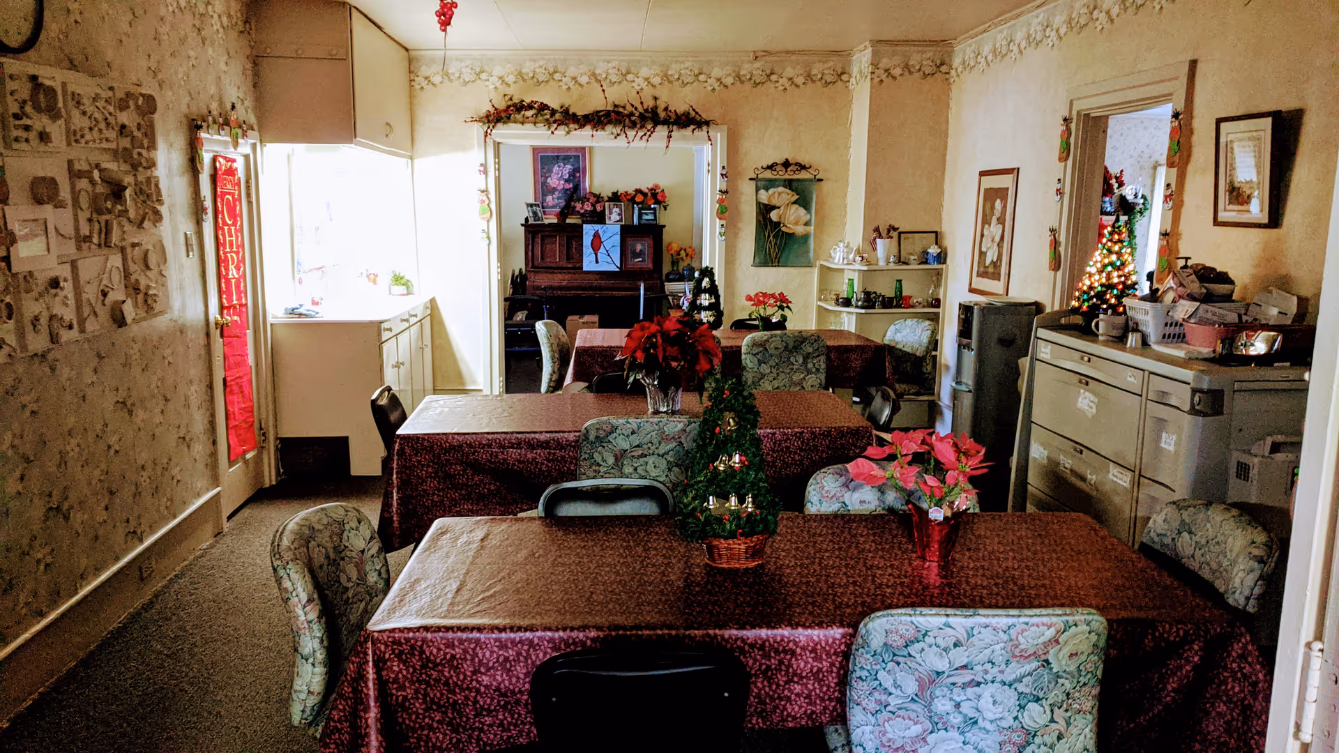 Interior dining room with multiple tables covered in maroon tablecloths, floral-upholstered chairs, holiday decorations, and a piano in the background.