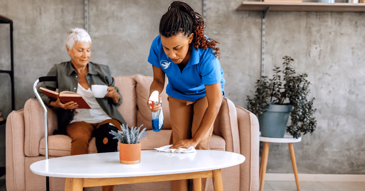 Caregiver cleans coffee table while resident sits on pink sofa reading nearby.
