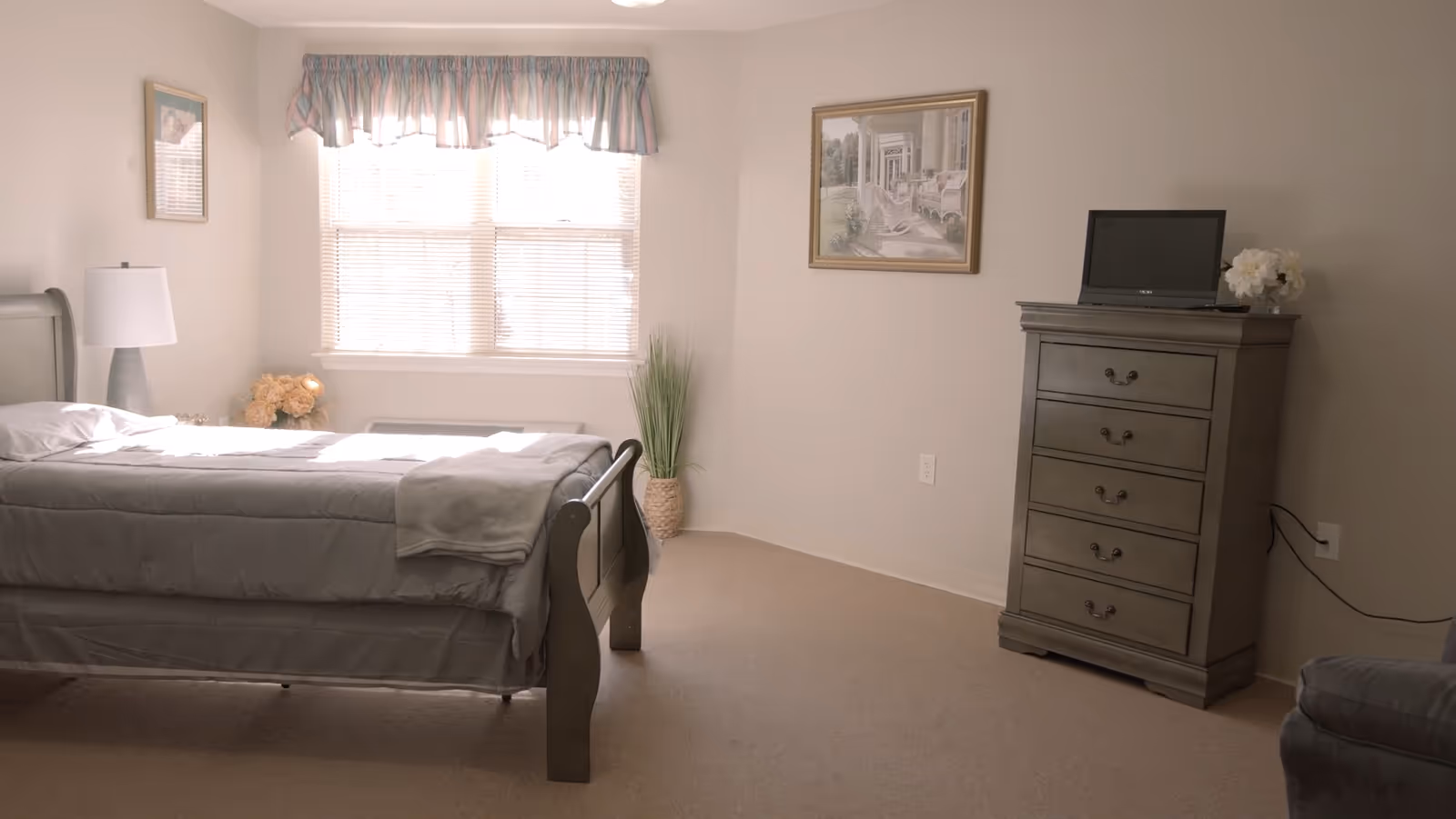 A bright bedroom with a large window covered by blinds and a pastel-colored valance. The room features a bed with gray bedding, a bedside lamp, a vase with flowers, a tall dresser with a small TV on top, and a framed picture on the wall.