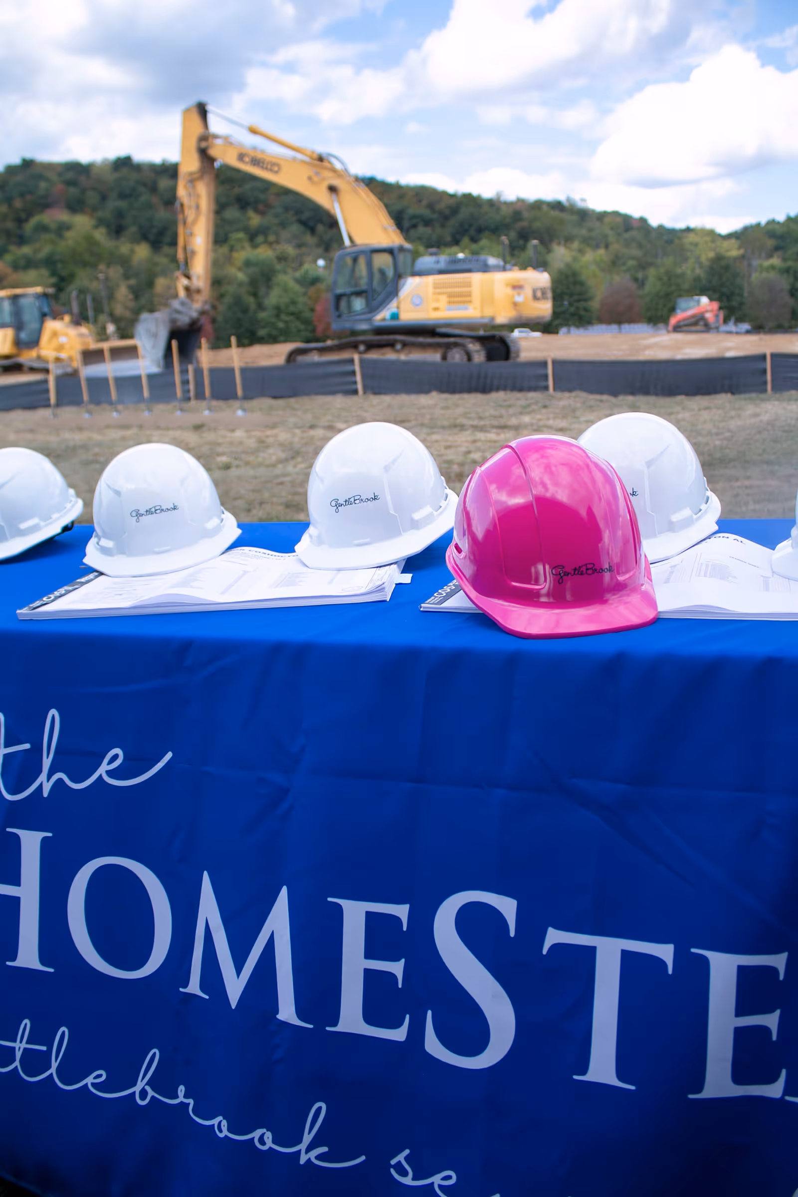 Outdoor construction site with an excavator behind a blue table banner for The HomeStead and helmets.