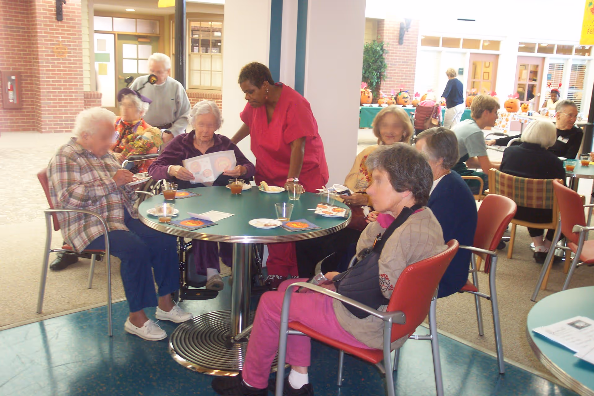 A group of elderly people sitting around tables in a communal indoor area, some eating and drinking, with a caregiver in red scrubs assisting one of the residents. The background shows more people and festive pumpkin decorations on a table.