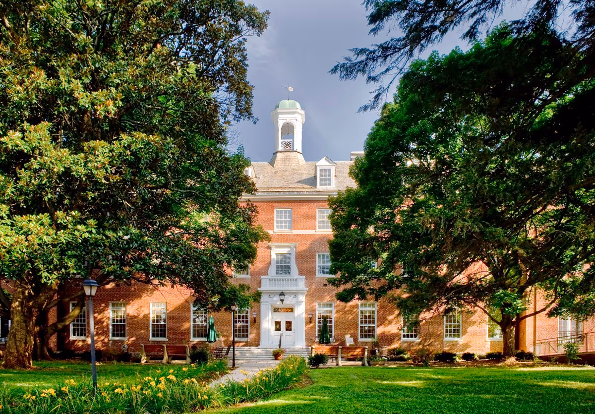 Brick senior living building with white entrance, green lawn, and surrounding trees.