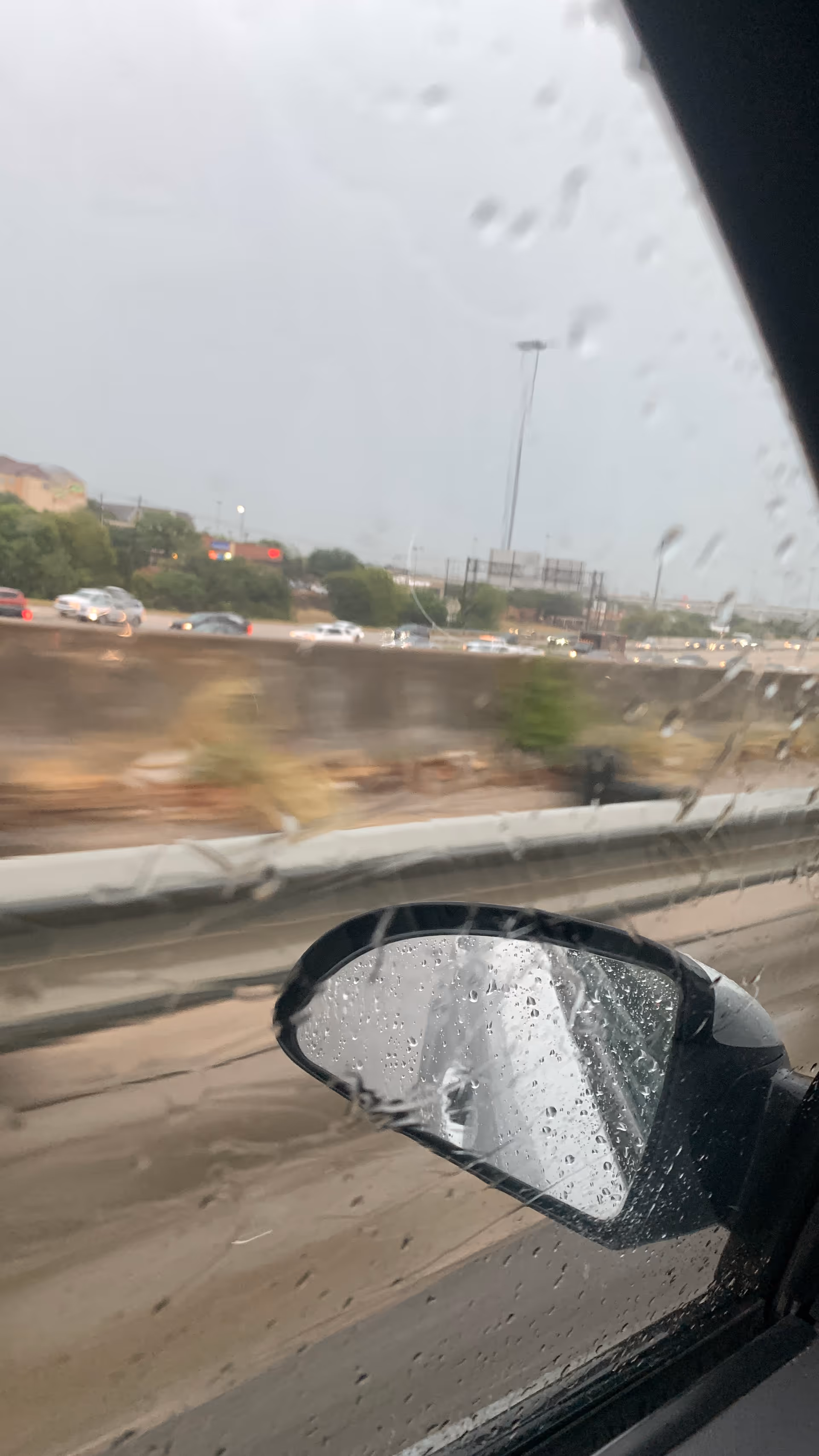 Rainy car window view of highway with guardrail and distant buildings; raindrops on glass.