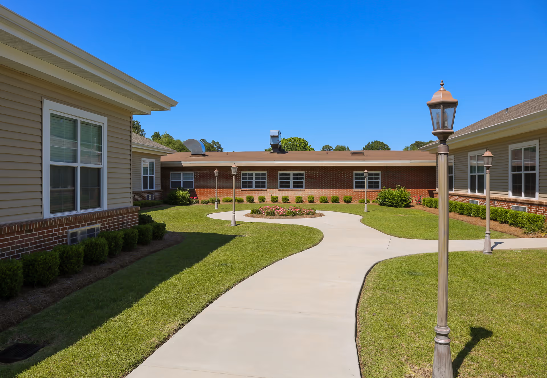 Sunny courtyard of a senior living facility with brick buildings, green lawn, and curved walkway
