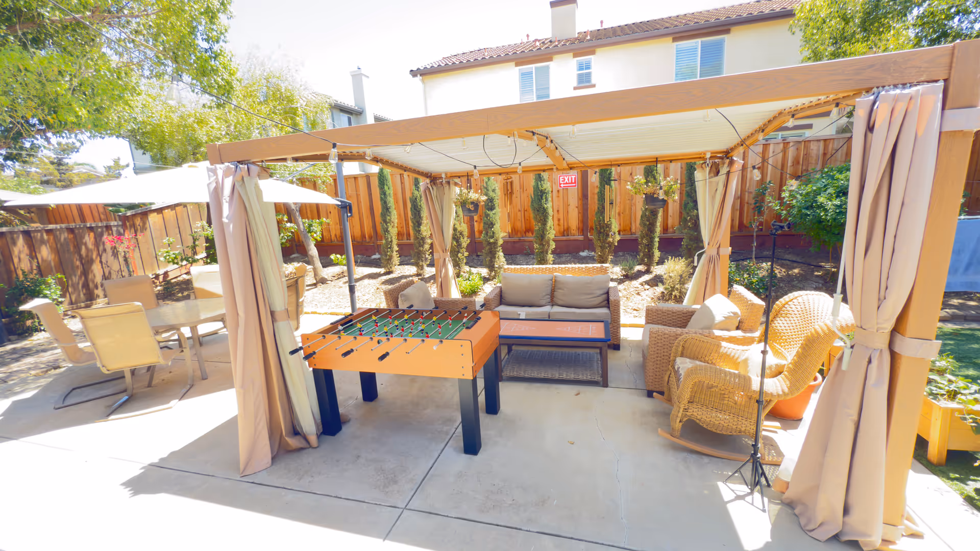 Outdoor seating area under a shaded pergola with wicker furniture and a foosball table.