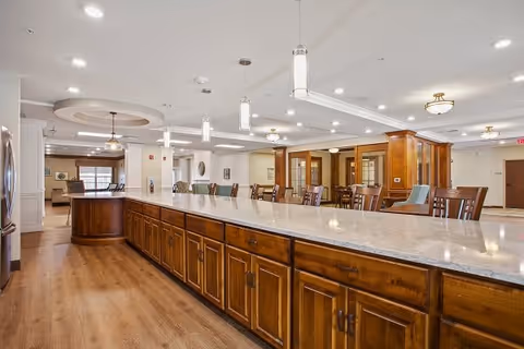 Communal dining area with a long marble-topped island, wooden cabinetry and multiple dining tables and chairs.