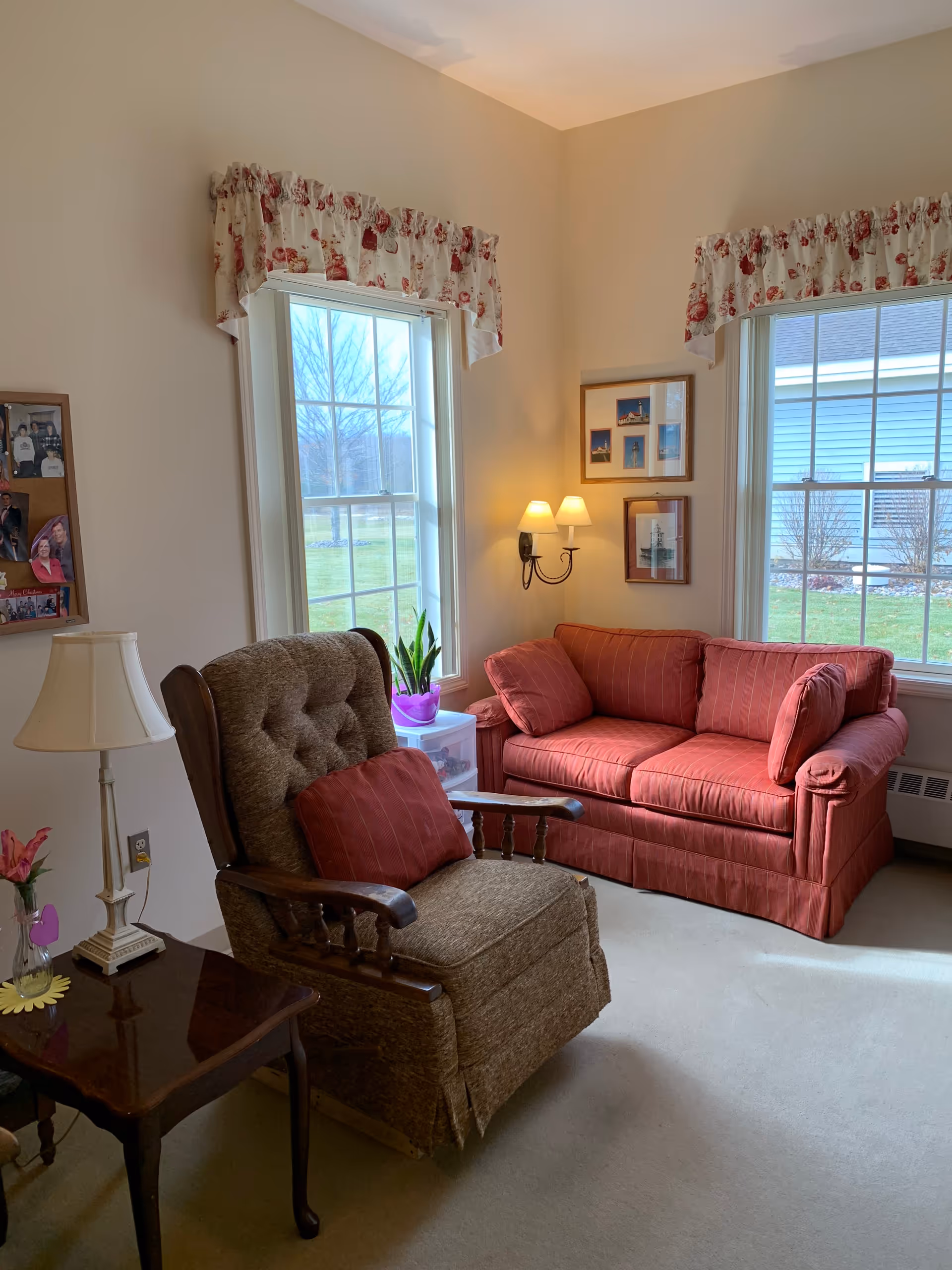 A cozy living room area with a brown upholstered armchair with a red cushion, a red striped loveseat with matching cushions, two large windows with floral valances, a small wooden side table with a lamp and a flower vase, and framed pictures on the wall.