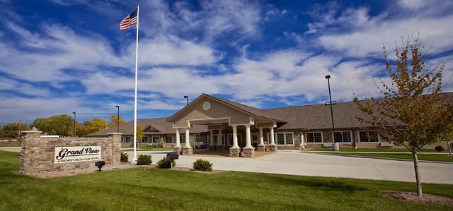 Exterior view of the Grand View Alzheimer's Special Care Center building with a covered entrance, a flagpole with an American flag, a stone sign displaying the facility name, and a well-maintained lawn under a partly cloudy blue sky.