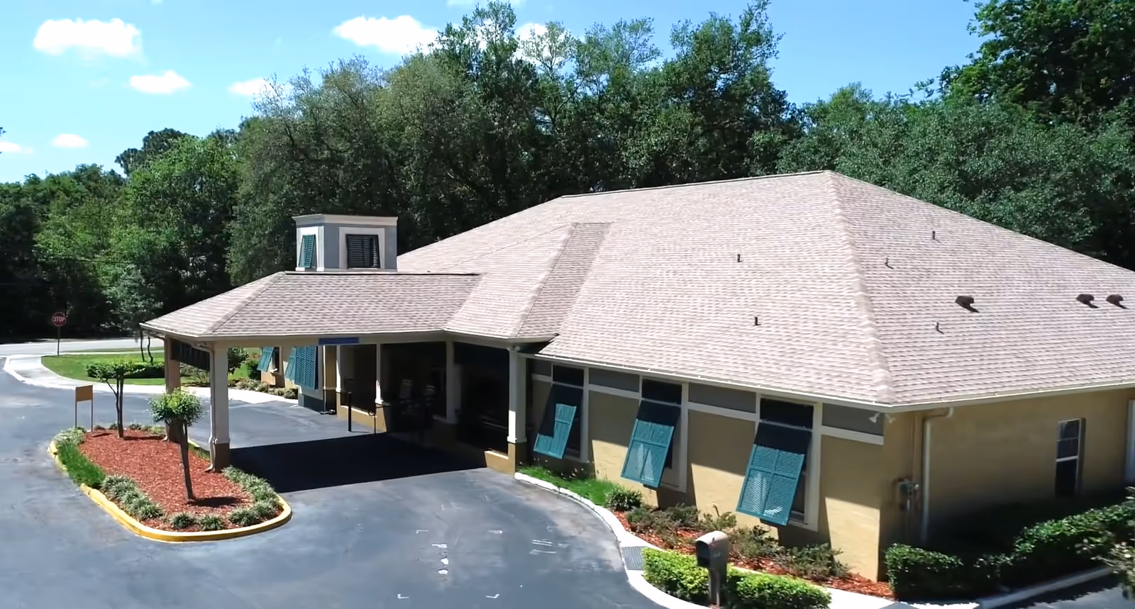 Front entrance and porte-cochère of a single-story memory care building with teal window shutters, surrounding landscaping, and trees in the background.