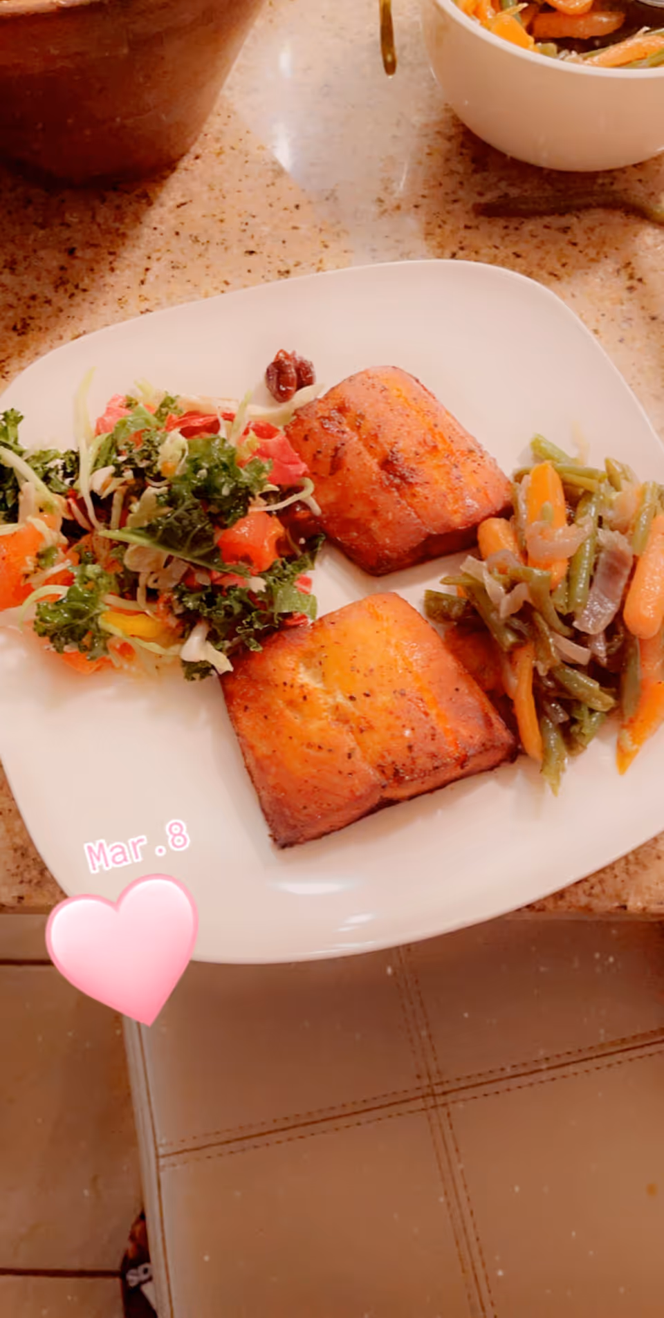 A white square plate with two pieces of grilled salmon, a serving of mixed sautéed vegetables including green beans and carrots, and a fresh salad with kale, tomatoes, and other greens on a speckled countertop.
