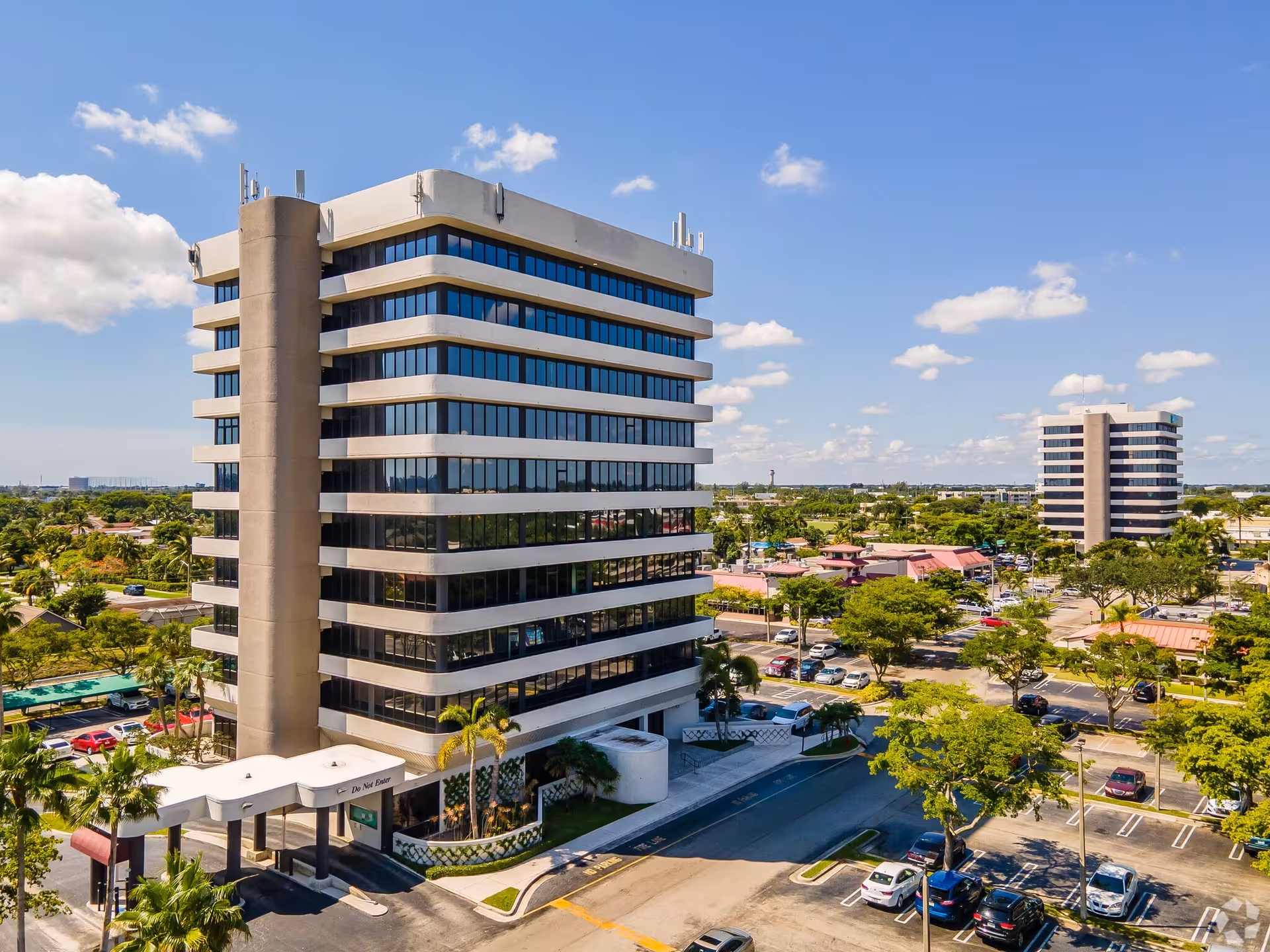 A tall modern building with glass windows, palm trees, and a surrounding parking lot.