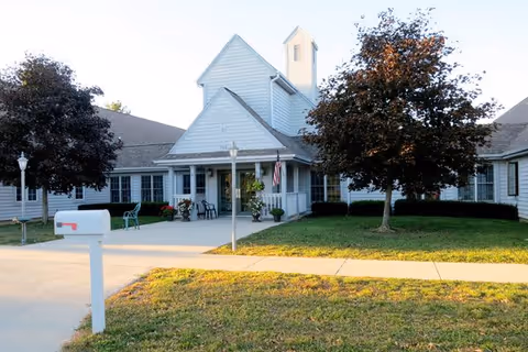 Front entrance of a single-story white senior living facility with a covered porch, mailbox, and landscaped lawn.
