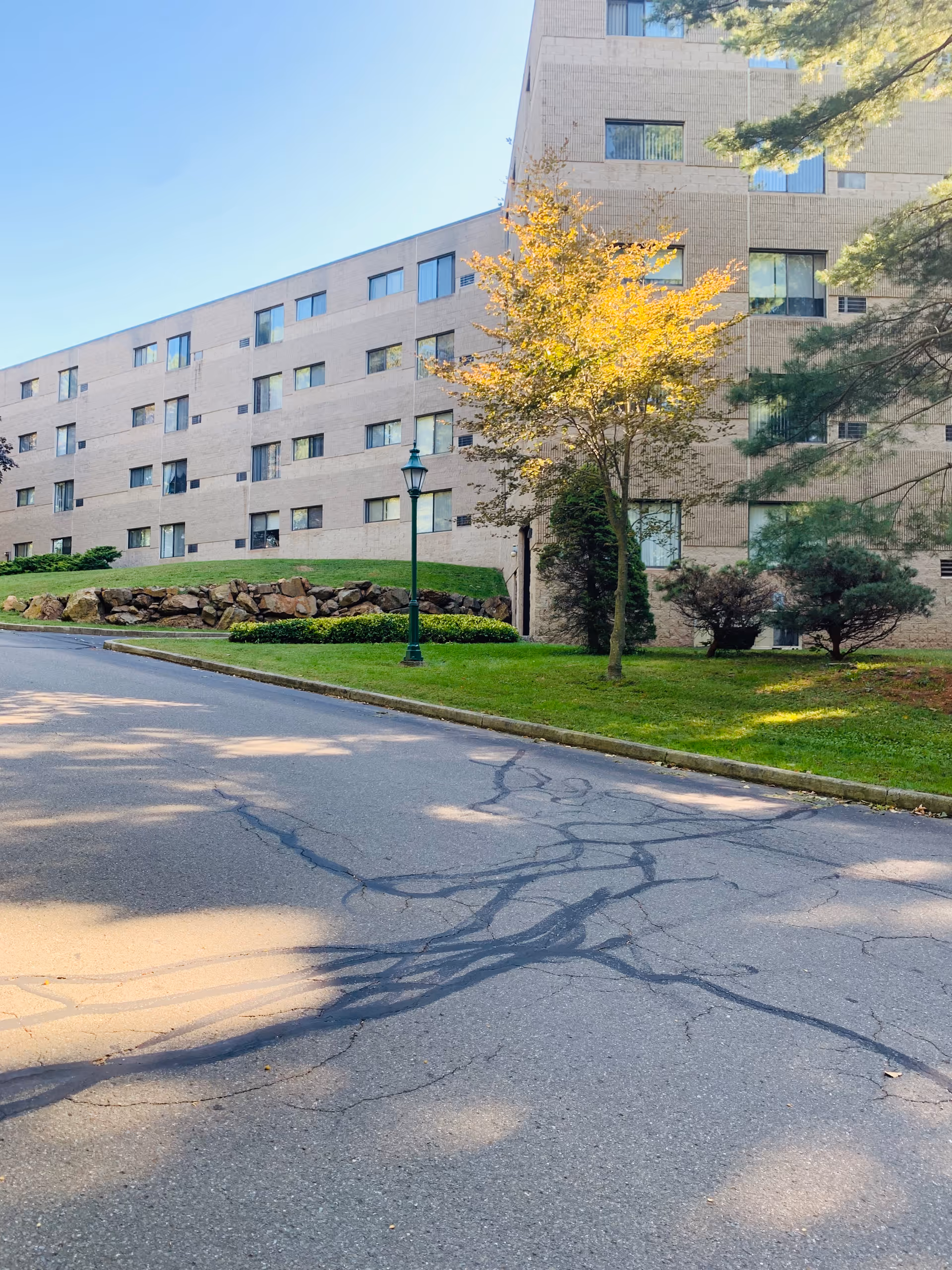 A multi-story brick senior housing building with a lawn, trees, and a street in the foreground.