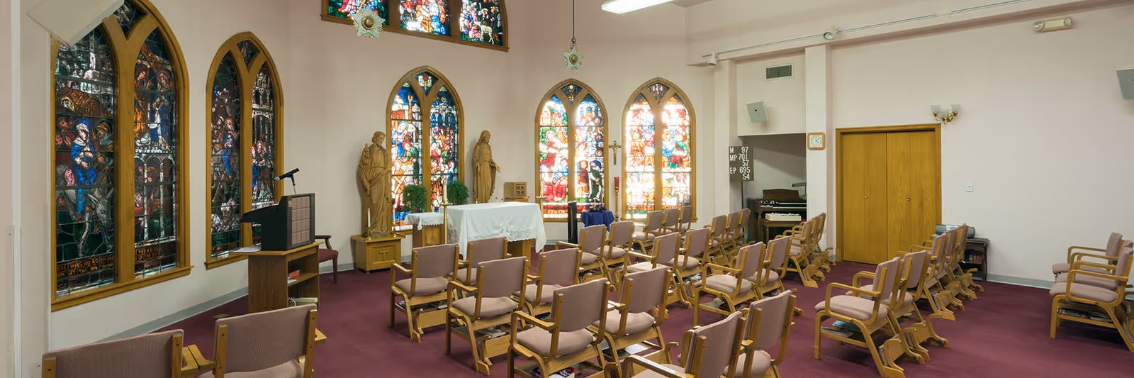 Interior chapel with rows of upholstered chairs facing an altar beneath stained-glass windows and religious statues.