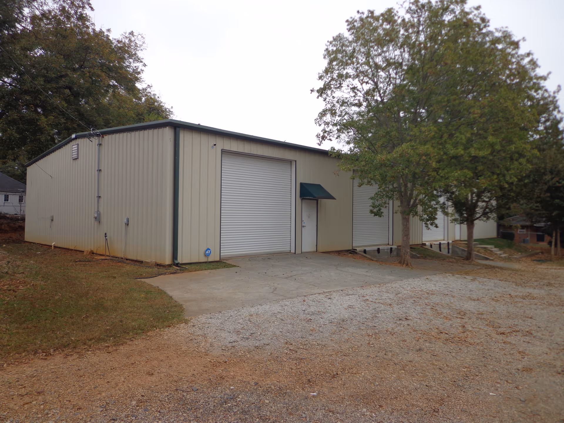 Beige metal building with large roll-up doors beside trees on a gravel lot.
