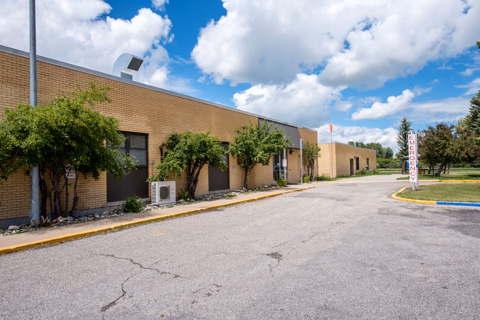 A single-story brick medical center exterior with trees, a parking lot, and an emergency sign.