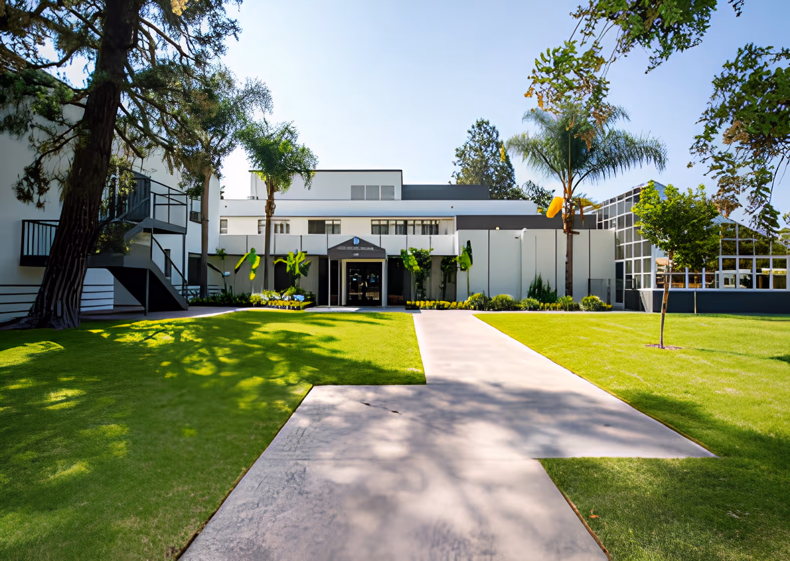 Front exterior view of French Park Care Center showing a modern white building with large windows, surrounded by green lawns, palm trees, and a clear blue sky.