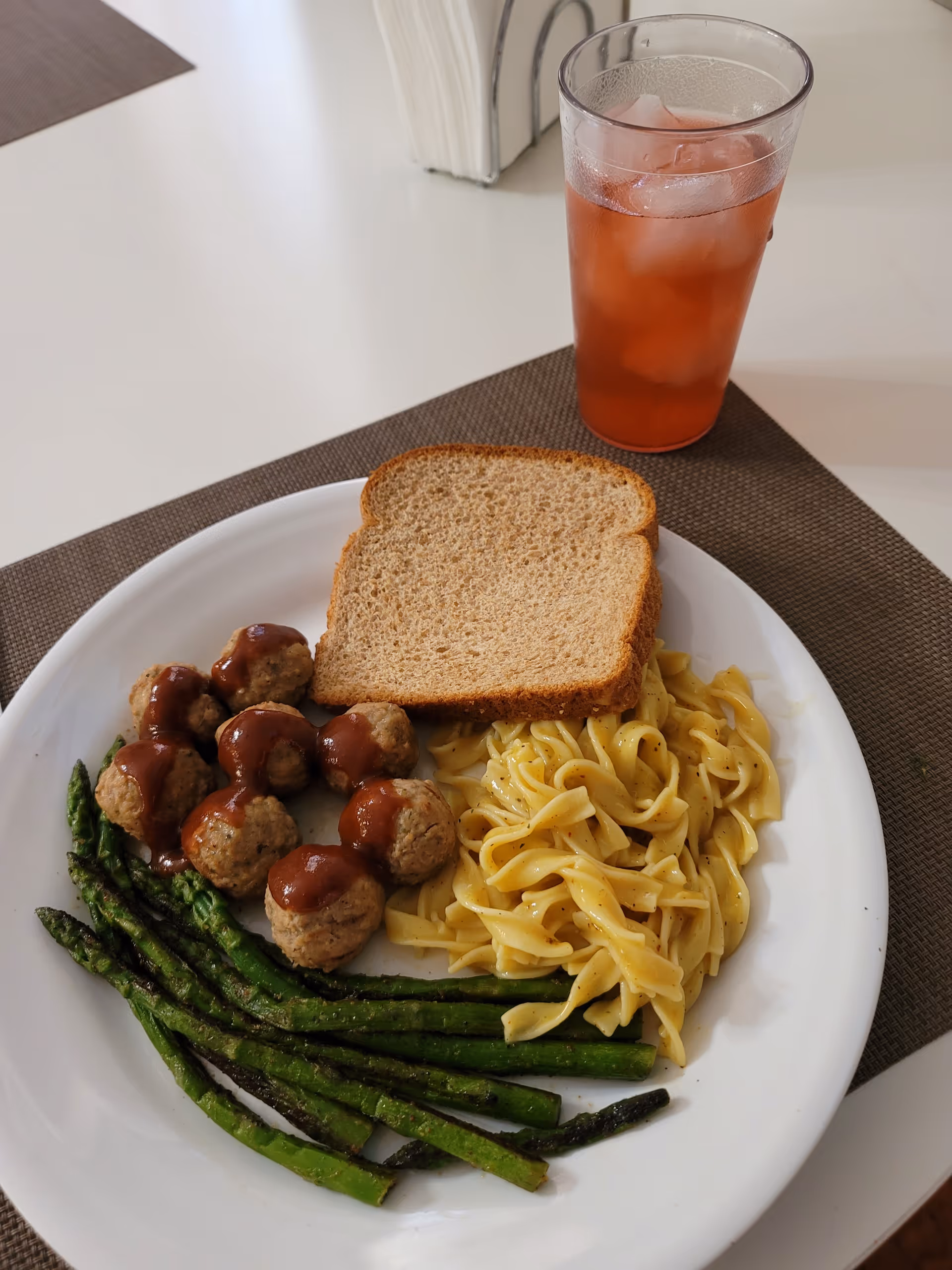 Plate with meatballs, pasta, asparagus, bread, and iced tea on a dining table.