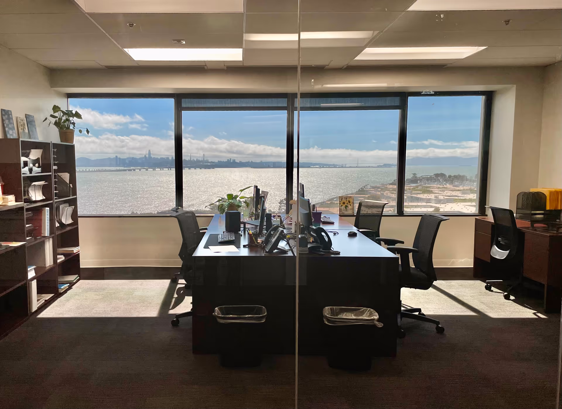Office workspace with desks, chairs, and a large window overlooking water and skyline.